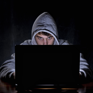 A hacker sitting at a laptop in a dark room, wearing a grey hoodie.